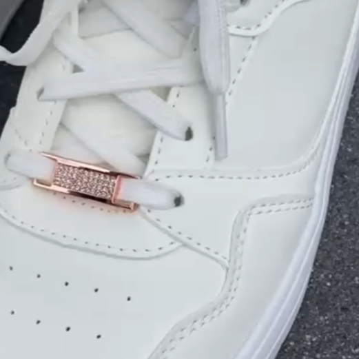 Nahaufnahme eines weißen Sneakers auf Asphalt; mittig an den Schnürsenkeln ist ein roségoldener Schuhcharm mit Kristallbesatz befestigt